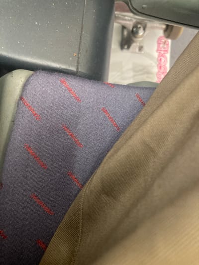 The image shows a close-up view of a seat on public transport. The seat is upholstered in a dark blue fabric with a repeating pattern of thin red lines. A person's light brown pants are visible in the foreground, partially obscuring the seat. In the background, through a gap in the seat structure, a white package with pink text is visible, possibly the packaging for a skateboard. The overall impression is one of a utilitarian interior, likely a bus or train. The image does not contain any visual information that would allow for the determination of its location in Montcada i Reixac, Spain.