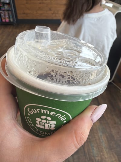 In Verin Ptghni, Armenia, a hand with light pink nail polish holds a green paper cup with a clear plastic lid. The lid has condensation on it and a small opening for drinking. The cup has a white rim and a logo with the text "Gurmenia Gastrom..." in white lettering and a graphic design. The background is blurred, showing wooden flooring and a person in a white shirt.