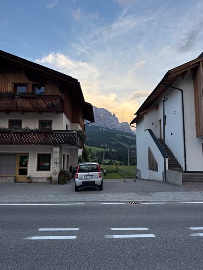 The image depicts a street view in La Villa, Italy, with two buildings flanking a path leading towards a mountainous landscape. The building on the left is a traditional wooden and stucco structure with balconies adorned with flower boxes. The building on the right is a modern white structure with an external staircase. A silver Fiat Panda is parked in front of the left building. In the background, rolling green hills rise to meet jagged, majestic mountains under a cloudy sky. The road in the foreground features white dashed lines and a drainage grate.
