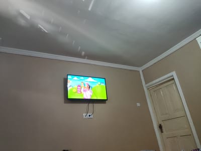 A television mounted on a light brown wall displays a cartoon scene of a woman in a pink outfit in front of colorful houses and trees under a blue sky with clouds. Below the television, a white electrical outlet is visible. To the right of the television, a light-colored, paneled door with a dark metal handle is set into the wall. The ceiling above is white and features decorative molding along the edges. The room is dimly lit, with light reflecting off the ceiling. The overall scene is consistent with a residential interior in Кипчок, Tajikistan.