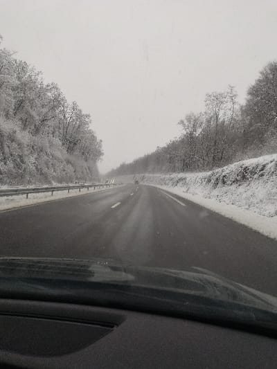 The image captures a winter scene on a multi-lane highway in Crusnes, France, under an overcast sky. The view is from inside a vehicle, with a dark dashboard and the lower portion of the windshield visible in the foreground. The asphalt road surface is dark and appears wet or slushy, marked with white lane lines. Both sides of the highway are bordered by snow-covered verges and embankments densely populated with trees and bushes, all heavily frosted with white snow. A metallic guardrail runs along the left shoulder of the road. In the mid-distance, several vehicles are observed traveling in the same direction, indicated by faint points of light from their illumination. Visibility is reduced by the prevailing snowy or hazy atmospheric conditions.
