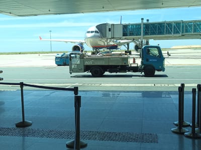 The image shows an airport tarmac scene at Cam Ranh International Airport in Bắc Cam Ranh, Vietnam. A large passenger airplane is parked at a gate, connected to a jet bridge. In the foreground, a blue service truck with "AGS AVIATION GROUND SERVICES CO., LTD" written on its side is positioned on the tarmac. The truck appears to be performing some type of service for the aircraft. Other airport vehicles and equipment are also visible. The scene is set under a clear blue sky with some clouds. The foreground shows the interior of an airport terminal with a tiled floor and stanchions with blue ropes, suggesting a waiting area.