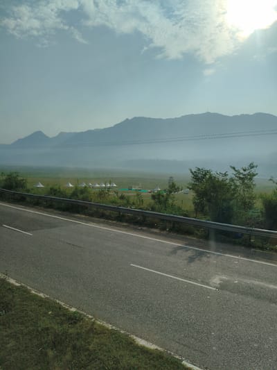 The image depicts a wide, paved road in the foreground, with a grassy shoulder to the left. The road appears to be a two-lane highway, with a white line marking the center and the edge. In the middle ground, there is a metal guardrail separating the road from a large, open field. The field is dotted with several conical white tents, suggesting a temporary encampment or event. Beyond the field, a line of lush green trees and vegetation creates a natural barrier. In the background, a hazy mountain range stretches across the horizon, with the sky above a pale blue, partially obscured by wispy white clouds. The sun is visible, casting a bright glow through the clouds. The overall scene suggests a rural or semi-rural setting, possibly on the outskirts of Bilaspur, India, given the presence of the highway and the distant mountains. There are no people visible in the image.