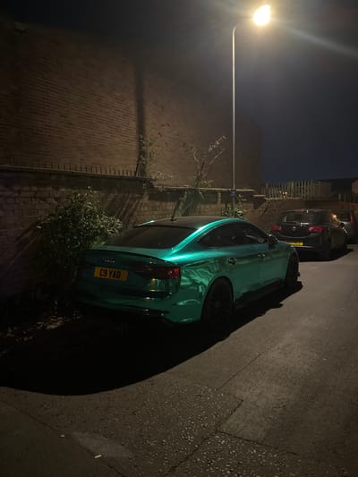 The image captures a nighttime scene on a street or alleyway in South Kesteven, United Kingdom. Two cars are parked along a paved surface next to a tall brick wall.
In the foreground, a prominent teal-colored car, identifiable as an Audi from the rear emblem, is parked, facing away from the viewer. Its rear license plate clearly displays "C9 YAD". The car has black multi-spoke wheels and a glossy finish that reflects the artificial light, creating bright highlights. Thick vegetation, resembling bushes or overgrown weeds, is visible around the car's base and along the brick wall to its left.
Behind the teal car, a second, darker-colored car (possibly black or dark grey) is also parked. Its rear license plate is partially visible, showing "FL71 EKL". The ground where the cars are parked appears to be asphalt or tarmac, transitioning to a patterned brick paving closer to the wall.
To the far left of the frame, a bright, glowing street lamp illuminates the scene, casting long shadows across the road and the adjacent brick wall. The sky above is dark, indicating the time of day is night. The brick wall features some vertical elements that resemble a small ladder or railing. No people are visible in the image. The scene suggests a quiet, urban or suburban setting after dark.
