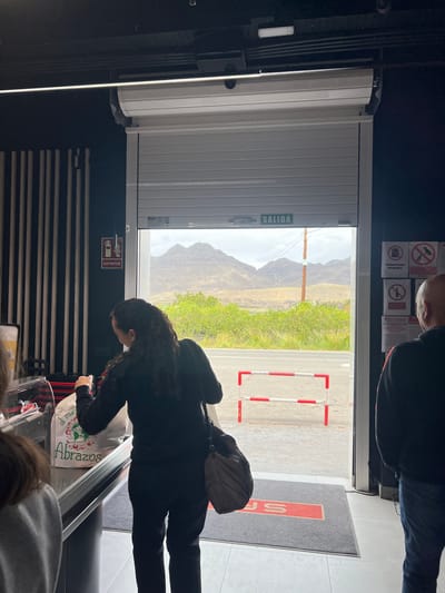 The image captures an indoor scene in La Aldea de San Nicolás, Spain. The focus is on a person in dark clothing leaning over a checkout counter. The person is holding a white shopping bag with the word "Abrazos" printed on it. In the background, a partially opened white roller shutter door reveals a view of mountains and green vegetation outside. A sign with the word "SALIDA" (Exit) is visible on the shutter door. On the wall, there are warning signs and a fire extinguisher. The flooring appears to be tiled. Other individuals are present, but partially out of frame.