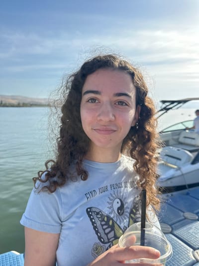 The image shows a young woman with curly brown hair standing on a dock. She is wearing a light blue t-shirt with a butterfly and the words "Find Your Peace of Mind" printed on it. She is holding a clear plastic cup with a black straw, and she is looking at the camera. The background shows a body of water, possibly the Sea of Galilee, and a docked boat. The coordinates provided (latitude: 32.844, longitude: 35.52) place the scene in Migdal, Israel.