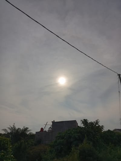 This image captures a cloudy sky with the sun visible as a bright, diffused circle. A power line stretches diagonally across the upper portion of the frame. Below the sky, lush green foliage is visible, with trees in the foreground. Behind the trees, a building under construction is partially obscured. The building appears to have at least two stories, with a red water tank on top. Construction materials and scaffolding are also visible. The scene suggests an outdoor setting in a somewhat developed area, possibly on the outskirts of a city like Kualu, Indonesia, given the presence of both natural vegetation and ongoing construction. The lighting indicates it could be daytime, though the cloud cover softens the sunlight. No people are visible in the image.