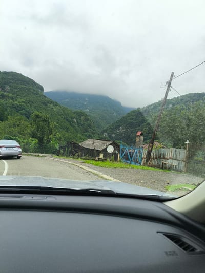 A silver sedan with license plate GQ 493 CC is parked on the side of a paved road, facing away from the camera. The car is on the left side of the image, positioned on the edge of the road where it curves. The road is asphalt with white lane markings. To the right of the road, a dirt and gravel area extends towards a collection of buildings.

The buildings appear to be in a state of disrepair. The closest building has a dark, sloped roof and appears to be constructed of wood and corrugated metal. Further back, a blue metal gate stands open, leading to a yard with a tall, brick structure resembling a chimney or tower with a red roof. Behind the gate, a corrugated metal fence encloses the property.

The scene is set against a backdrop of lush, green mountains. The slopes are covered in dense vegetation, with some rocky outcrops visible. The sky is overcast with heavy, gray clouds, suggesting it may be raining or has recently rained, as evidenced by a few raindrops visible on the windshield from which the photo is taken. A wooden utility pole with wires runs diagonally across the upper right portion of the image, near the mountainside.

The interior of a vehicle is visible in the foreground, with the dashboard and windshield occupying the lower part of the frame. The environment suggests a rural or semi-rural setting, likely in a mountainous region.