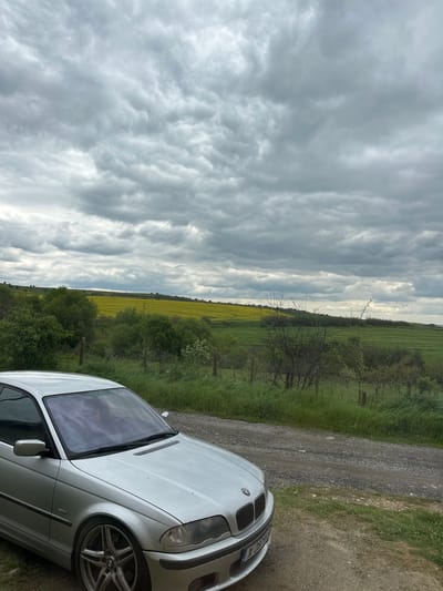 The image shows a silver BMW 3 Series parked on a gravel road next to a green field in Rogozinovo, Bulgaria. The sky is overcast with dense, gray clouds. The field is predominantly green, with a strip of yellow flowers running through it. A wire fence borders the field. There are no people visible in the frame. The car is positioned at an angle, with its front end facing the viewer.