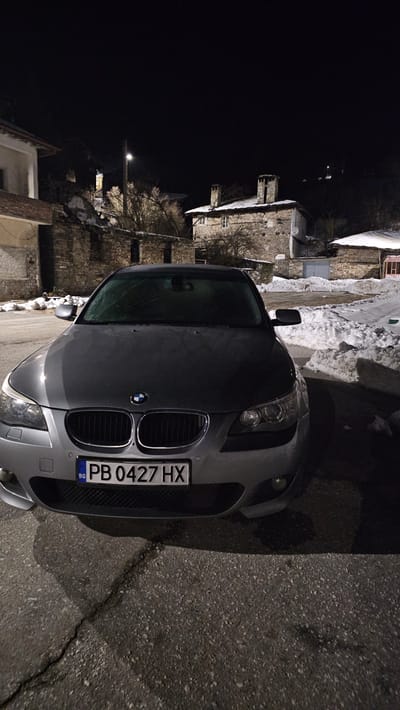 The image depicts a static scene at night in the village of Sitovo, Bulgaria, during winter. A dark grey BMW 5-Series sedan, model E60/E61, is parked on an asphalt surface, positioned slightly angled towards the left, with its front facing the viewer. The windshield of the vehicle is frosted or covered with condensation. The front license plate, "PB 0427 HX", is clearly visible, identifying the vehicle as registered in the Plovdiv region of Bulgaria.
In the background, traditional stone architecture is evident. To the left, a partial view of a light-colored building with a dark roofline is visible, alongside what appears to be a stone ruin or partially collapsed structure. Centered in the mid-ground is a two-story stone house with a snow-covered tiled roof and two prominent stone chimneys. Two windows are visible on its facade. To the right of this house, another smaller stone building with a snow-covered roof is present. Bare trees and shrubs are scattered throughout the background, indicative of winter conditions.
Snow has accumulated on the roofs of the buildings and on the ground, forming drifts along the sides of the asphalt surface, particularly to the right of the parked vehicle. Illumination is provided by a single streetlamp mounted on a pole, visible in the background, which casts light on portions of the trees and buildings. The sky is completely dark, indicating nighttime. No individuals are present in the scene, and no specific actions or interactions are depicted. The asphalt ground in the foreground shows minor cracks.