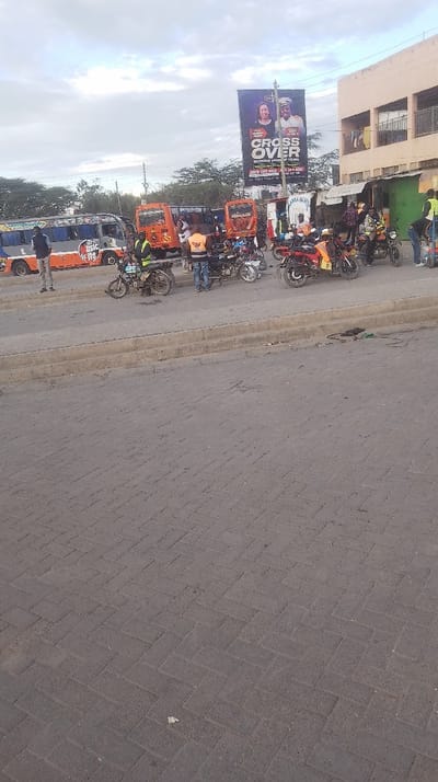 A busy street scene in a developing country. Several motorcycles and three small orange and blue buses are parked along the side of the road. People stand near the vehicles, some wearing orange vests. A large billboard advertising a "Crossover Service" is visible in the background. A two-story building is situated near the billboard. The road surface is paved with concrete bricks, and the sky is overcast.