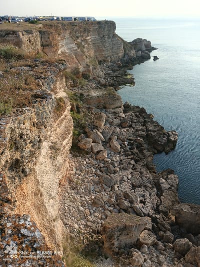 The image depicts a rugged cliffside overlooking a calm sea. The cliffs are composed of layered sedimentary rock, with visible strata and some areas of erosion. Sparse vegetation, including dry grasses and a few small purple flowers, grows on the cliff tops and ledges. Below the cliffs, a jumble of large rocks and boulders forms a rocky shoreline. The sea is a deep blue with gentle ripples. In the distance, the horizon is clear, with a pale sky. On the very top of the cliff in the background, several recreational vehicles, tents, and cars are parked, suggesting a campsite or a public gathering area. The overall scene is natural and serene, with human presence limited to the distant vehicles. The image captures the natural beauty of the coastline in Kamen bryag, Bulgaria, known for its dramatic cliffs and clear waters.