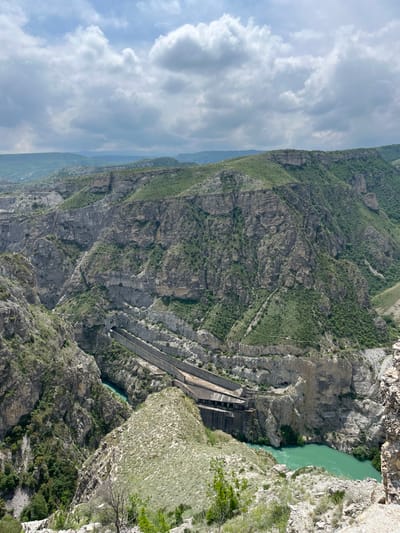 The image depicts a landscape with a notable hydroelectric dam feature. Located in Chirkey, Russia, the scene presents a deep gorge or canyon carved by a river flowing through it, exhibiting a turquoise color. The dam structure is visible, constructed of gray concrete and extending across the gorge. The steep canyon walls show rocky terrain with sparse vegetation. The sky above is partly cloudy, suggesting fair weather. The overall perspective is from a high vantage point looking downward. There are no discernible people in the image.