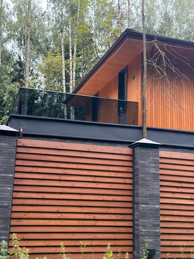 A modern wooden house with a glass balcony is visible behind a horizontally slatted wooden fence with dark gray brick posts in Новодарьино Ран Дп, Russia. The house has a pitched wooden roof and vertical wooden siding. A bare tree branch extends from the right side of the frame, touching the side of the house. The environment is wooded with tall trees visible behind the house. The fence has a dark gray metal beam above it, and the brick posts are capped with dark gray pyramid-shaped finials. Green foliage grows at the bottom of the frame in front of the fence.