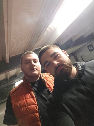 A digital image depicts two adult males posing for a selfie in an indoor parking garage, identified as being in Halle (Saale), Germany. The environment is characterized by a low ceiling with light-colored corrugated panels and multiple exposed fluorescent light fixtures providing overhead illumination. In the upper right background, a white sign with the black alphanumeric designation "P6" is visible on a darker wall surface.

The male subject on the left is lighter-skinned, with short-cropped light hair and a light beard. He is wearing a black t-shirt beneath an unzipped orange puffer vest. His gaze is directed generally towards the camera.

The male subject on the right, positioned closer to the camera, is darker-skinned with short, dark hair and a full dark beard. He is wearing a dark, possibly black, zippered hooded jacket, with a silver-colored chain visible around his neck. His head is slightly tilted, and he is looking directly into the camera. Both individuals are standing static, posing for the photograph, with no active interactions or discernible actions other than posing.
