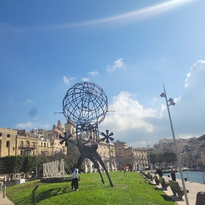 An outdoor, daytime scene at Xatt ir-Risq, Cospicua, Malta, captures a panoramic view under a partly cloudy blue sky with bright sunlight. In the center-left foreground, a large, dark-colored metal sculpture depicting a spherical globe with a grid-like structure rests on an abstract, multi-legged base featuring propeller-like elements. This sculpture stands on a manicured grassy mound. To its immediate left, a white sign displaying the partial text "Bormla" is visible. Two individuals are near the sculpture on the grass; one person in a white top and dark trousers stands facing away from the camera.
To the right, a paved promenade extends alongside a body of water, likely a part of the Grand Harbour. Multiple dark-colored benches with wooden slats are arranged along the promenade, facing the water. Several dark mooring bollards are positioned along the quay edge. Three additional individuals are visible further down the promenade to the right; one appears to be bending over. A tall, black lamppost with two directional lights stands prominently on the far right.
The background is dominated by traditional Maltese architecture: a continuous line of multi-story, light-colored stone buildings with balconies and arched windows. A prominent church with a large central dome and two smaller domes/bell towers rises above the buildings in the central background. Sparse trees are interspersed along the waterfront and among the buildings. The upper half of the image features a bright blue sky with scattered white cumulus clouds and a distinct sun flare in the upper right quadrant.