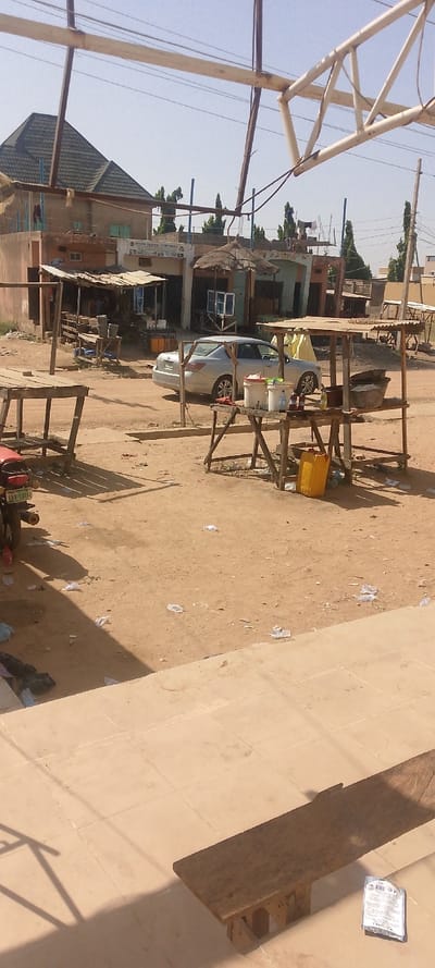 The image depicts an unpaved outdoor market area in Danbare, Nigeria, under a clear sky. In the foreground, light-colored tiled ground features a narrow wooden bench and a small white packet. The midground consists of a dusty, unpaved surface strewn with scattered plastic debris. Multiple wooden market stalls are visible: a bare frame on the left, a stall with a corrugated metal roof, and a stall on the right holding white buckets, bottles, a metal basin, and a yellow plastic jerrycan. A silver Honda sedan is parked in the midground, with a person in a yellow garment partially visible behind it. A red motorcycle with license plate AKR-530JQ is partially visible on the far left. The background comprises a row of single and two-story buildings, including one with a dark pitched roof and another displaying a sign that reads "MISAN CENTRAL VENTURES." Tall green trees, utility poles, and overhead power lines are visible against the clear blue sky.