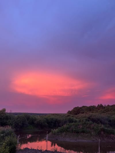 The image depicts a serene natural landscape at dusk, with a vibrant sky painted in hues of purple, pink, and orange. A body of water, possibly a river or lake, occupies the lower portion of the frame, reflecting the colorful sky. Lush green vegetation, including trees and bushes, lines the banks of the water and stretches towards the horizon. The sky is dominated by a large, illuminated cloud formation that glows with intense pink and orange light, contrasting with the deeper purple and blue tones of the upper sky. The overall scene suggests a tranquil and picturesque natural setting, likely in a rural or wilderness area. No people or specific urban features are visible in the image.