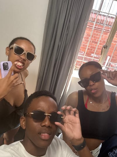 This media file is a mirror selfie featuring three young adults, likely taken indoors during the daytime, suggested by the bright natural light. The setting appears to be a room with a window, offering a glimpse of an exterior brick wall and white security bars. Gray curtains or blinds are drawn across a section of the background. The location context provided is Port Alfred, South Africa.

The subjects are posing playfully, all wearing sunglasses. The person in the top-left section of the image wears a black tank top with a red bra strap visible and a delicate necklace. They have dark, rectangular sunglasses and are sticking their tongue out slightly. The individual in the bottom-right portion wears a white t-shirt and rimless black sunglasses with gold accents, also seen with their tongue out. A black smartwatch is visible on their wrist. The third person, positioned in the bottom-left, wears a light-colored top and large, light-colored cat-eye sunglasses. They are captured in a wide grin, showing their teeth, while holding a light purple smartphone, which is presumably being used to take the picture.

The overall atmosphere of the image is casual and joyful, with the subjects exhibiting playful expressions. No discernible text is visible within the image.