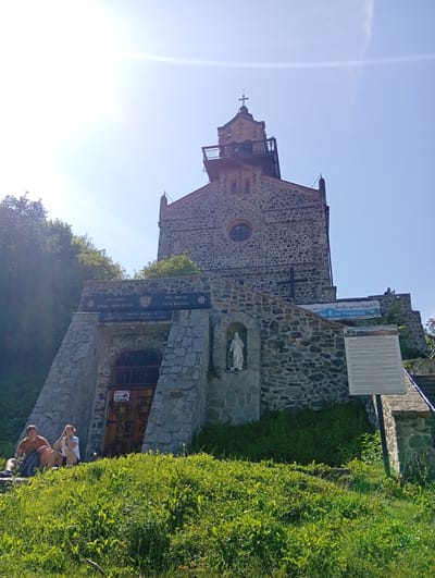 The image shows a stone church atop a hill in Sobótka, Poland. The church has a tower with a cross on top and a balcony. The facade of the church is made of stone, with a circular window and a large wooden door. There are two people sitting on the grass in front of the church, and a statue of a woman in a niche on the side of the building. A sign in Polish and English provides information about the history of the church and the mountain. The sky is clear and blue, and the sun is shining. The church appears to be a popular tourist destination.