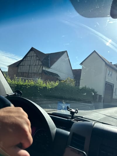 This media file is a first-person perspective shot from inside a vehicle, looking forward through the windshield. The image is taken on a clear, sunny day, suggesting it is daytime, likely mid-morning or afternoon given the bright sunlight and clear blue sky. The scene outside depicts a residential area with several houses visible. One house has a light-colored facade and a dark tiled roof, with visible window frames. Another building appears to be older, with a half-timbered upper section and a red brick lower section. A row of green bushes runs along the side of one of the buildings.
Inside the vehicle, a hand is visible gripping the steering wheel, indicating the person is driving. A black smartphone mount is attached to the windshield near the center of the frame. A portion of the car's dashboard, including the steering wheel and part of the instrument cluster, is also visible on the right side. The instrument cluster shows a dial with markings and numbers, suggesting it is a speedometer, with a portion of a digital display that appears to be pink.
The view through the windshield includes some smudges and reflections, characteristic of a car window. The overall mood of the image is bright and sunny, suggesting a pleasant day for driving. The location is identified as Volpertshausen, Germany. There is a small white sticker with blue text visible on the lower left side of the windshield, but the text is not legible.