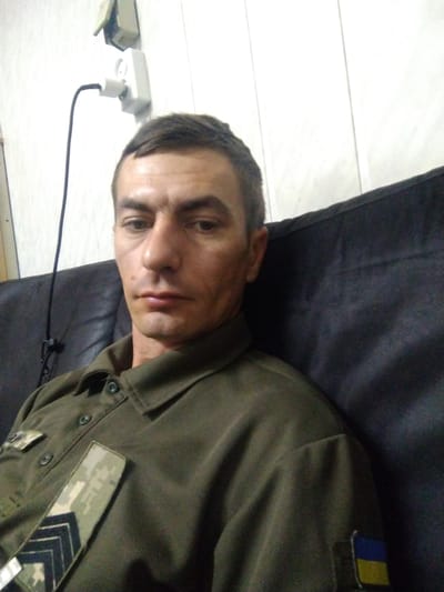 A man lies on a dark surface, possibly a couch or bed. He is wearing a military-style olive green uniform with patches on the left chest. One patch is a camouflage pattern with diagonal stripes, and another appears to be a flag of Ukraine. The man's head is turned towards the camera, and he is looking directly at it with a neutral expression. His face is illuminated by a light source that casts shadows, suggesting it may be daytime.

In the background, a white electrical outlet with a black cord plugged into it is visible on a white, horizontally-lined surface, which could be a floor or a table. A small, light-colored object, possibly a power adapter, is next to the outlet. The scene is dimly lit, and the overall impression is informal. The presence of military uniform and the Ukrainian flag patch suggests a connection to Ukraine, and the location context confirms Vylok, Ukraine. No specific time of day or weather conditions are discernible.