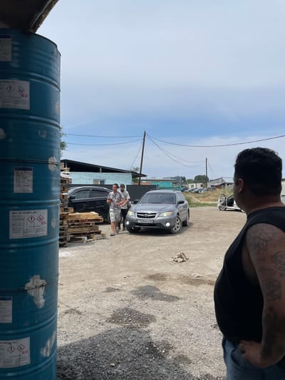 A man with tattoos on his arm and shoulder faces away from the camera, looking towards a courtyard in Кенен Әзірбаев ауылы, Kazakhstan. In the courtyard, two men walk past a stack of wooden pallets and a gray SUV. The SUV has the license plate "729BFG02". A black car is parked behind the SUV. A large blue drum with hazard labels is in the foreground to the left, partially obscuring the view. The scene is dusty with scattered gravel and rocks. In the background, there are buildings, power lines, and a utility pole under a cloudy sky.