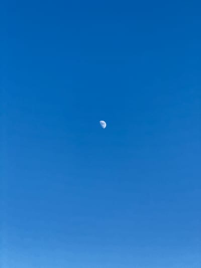 A photograph captures a clear, bright blue sky with a single, small, crescent moon visible. The moon is situated in the lower central portion of the frame. There are no people or other objects in the image, and the scene is entirely focused on the expanse of the sky. The lighting suggests it is daytime, as the sky is a vibrant blue and the moon is illuminated. The smooth, consistent color of the sky indicates a cloudless day. There are no discernable location cues to suggest it is specifically Mastaga, Azerbaijan. No text is visible in the image.