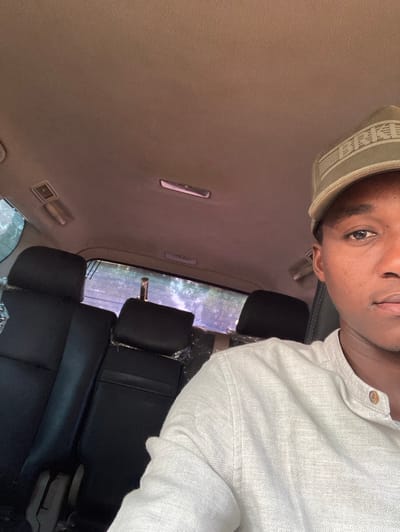 The image depicts a male individual inside a vehicle in Yan Dala, Nigeria. The individual, positioned to the right of the frame, is visible from the chest up, wearing a light-colored, possibly off-white or light grey, long-sleeved shirt with a mandarin collar and two light-brown buttons. He is also wearing an olive green baseball cap with a textured pattern on the front. His right eye and part of his face are visible, and he appears to be looking towards the camera. The vehicle's interior features a light brown textured headliner with visible overhead lights and vents. Behind the individual, two rows of unoccupied black or dark grey passenger seats are visible, with some seats, particularly in the middle row, covered in clear protective plastic. Through the vehicle's rear window, outdoor foliage is discernible under daylight conditions.
