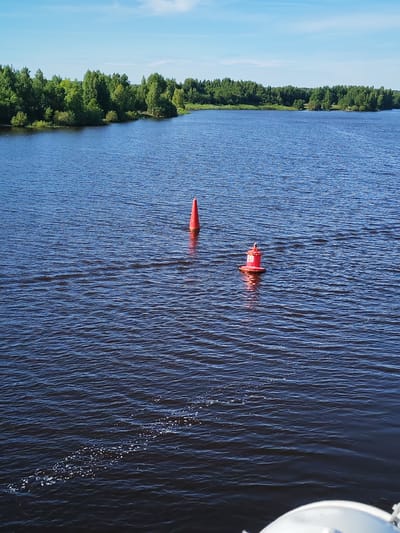 The image depicts a serene river scene in Nova, Russia, under a clear blue sky. The water, a deep blue, is rippled, suggesting a gentle breeze. Two red buoys are visible in the water, marking a channel or hazard. One buoy is a tall, conical shape, while the other is shorter and more cylindrical, with a white band and the number "20" clearly visible in black.
The shoreline on the far side of the river is lined with lush green trees, indicating a natural, undeveloped area. The foliage is dense and vibrant, suggesting it is likely summer. The light is bright, consistent with daytime, and there are no discernible clouds in the sky.
In the foreground, a portion of a white object, possibly the railing or part of a boat, is visible at the bottom right corner, implying the photo was taken from a vessel on the water. The overall atmosphere is peaceful and tranquil, capturing a moment of natural beauty. No people are visible in the image, and the focus is on the landscape and the buoys. The time of day appears to be mid-morning or early afternoon due to the quality of the sunlight.