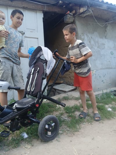 This media file, captured in Pishchanka, Ukraine, shows two young boys and a baby outdoors. One boy, wearing a striped t-shirt and red shorts, stands near a black tricycle. He appears to be interacting with the tricycle, possibly pushing it. The second boy, in a patterned t-shirt and camouflage shorts, is closer to the foreground, looking towards the camera. He is holding a baby, who is dressed in a yellow and white striped outfit.

The scene appears to be a casual, outdoor setting, likely a yard or alleyway, with a weathered building in the background and patchy grass and dirt on the ground. The lighting suggests it is daytime. The overall mood seems relaxed and familial. There is no visible text in the image.