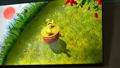 The image displays a digital rendering of a whimsical scene, possibly from an animated film or video game. The primary subject is a small, yellow, toy-like figure resembling a duck or chick, perched atop a decorative, cylindrical object. This object is predominantly red with intricate golden patterns and appears to be a music box or a similar trinket. The toy figure itself is detailed with brown markings on its head and body, and it has a proud stance.
The setting is a vibrant, stylized outdoor environment. The ground is covered in a thick, abstract representation of green grass, with streaks of yellow and gold suggesting sunlight filtering through. In the background, on the left side, a large, soft-edged red orb hangs in a pale, sky-like area, possibly representing a sun or a moon. Delicate, light-colored plant silhouettes are visible near this orb, adding to the ethereal atmosphere. Along the top right edge of the frame, there are clusters of bright red, berry-like objects hanging from thin stalks, adding a pop of contrasting color.
The overall mood of the image is cheerful and playful, evoking a sense of innocence and fantasy. The lighting suggests it might be daytime, perhaps during a sunrise or sunset, given the soft glow of the red orb and the warm tones of the grass. There are no discernible people or animals other than the toy figure. No specific actions are taking place beyond the figure being stationary on its decorative base. The location cues are not specific to Yakkabog, Uzbekistan, and are more characteristic of a fantastical, animated world. No visible text is present within the main image, although a small logo, possibly a brand name, is visible in the bottom right corner of the television screen frame, appearing as "meonk."