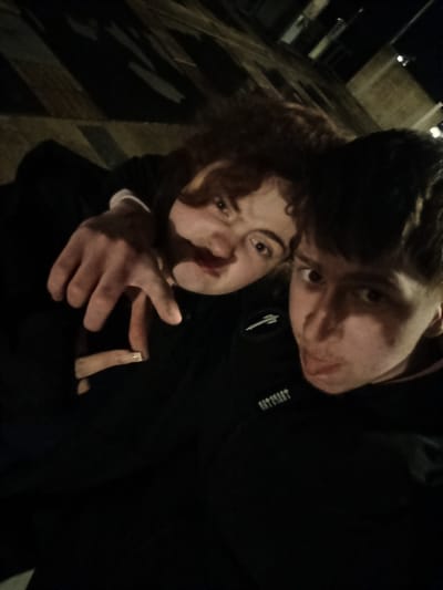 Two young people are posing for a selfie outdoors at night. The location is implied to be Emmerich on the Rhine, Germany, but this is based on contextual information not directly visible in the image. The person on the left has light skin, curly brown hair, and is making a hand gesture. The person on the right is closer to the camera, has short dark hair, light skin, and is sticking out their tongue. They are both wearing dark-colored clothing. In the background, there is a brick wall and some streetlight. The lighting is dim.