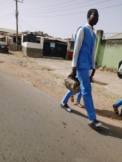 The image depicts an outdoor street scene in the vicinity of Kungulu School, Zambia, under a clear sky. In the foreground, a young male, likely a student, walks from right to left along an asphalt road. He wears a blue sleeveless vest over a white long-sleeved shirt, blue trousers, and black and white sandals over white socks. He carries a light-brown, patterned, rectangular lunchbox or small bag in his right hand. Behind him, partially obscured, are two additional individuals also appearing to wear similar blue uniforms and sandals, walking in the same direction.
To the right of the road is an unpaved dirt shoulder leading to a low, light-colored wall. To the left of the road, a dirt area extends towards a series of single-story or low-rise buildings. These structures feature painted walls (some white and grey), dark metal gates, and corrugated roofs. Two large black water tanks and several satellite dishes are visible on or adjacent to some buildings. A dark sedan is partially visible on the far left, parked near a building. A wooden utility pole with multiple overhead lines stands on the left side of the frame. The distant background reveals uneven, possibly rocky, terrain.
