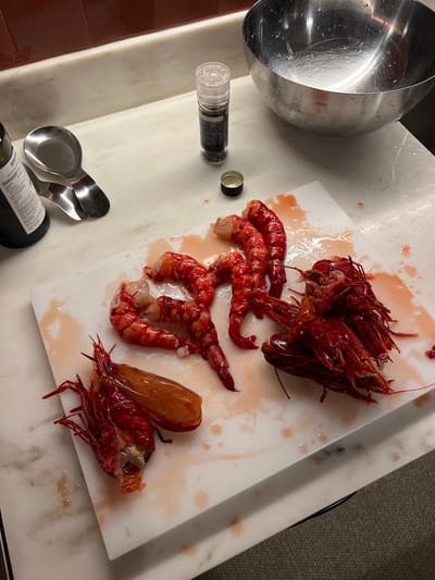 On a marble countertop in Gondarém, Portugal, sits a white cutting board. On the board are several raw, red prawns, some whole and some with the bodies separated from the heads and shells. The prawns are surrounded by a reddish liquid, possibly prawn juices. Next to the board is a metal mixing bowl, a pepper grinder, a black bottle with white lettering, and two metal spoons. The background includes a dark wood cabinet and a beige carpet.