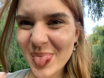In an outdoor setting, a young woman with light brown hair is captured in a close-up, front-facing photograph. Her eyes are open, and she is sticking her tongue out slightly. She is wearing a hoop earring with a pink heart-shaped charm. Her skin appears fair, with a small mole visible on her chin. The background consists of lush green foliage and trees, suggesting a natural, possibly park-like environment. The lighting indicates it is daytime, with sunlight filtering through the leaves. The image does not contain any recognizable landmarks or features that would allow for identification of a specific location within Derevky, Ukraine.