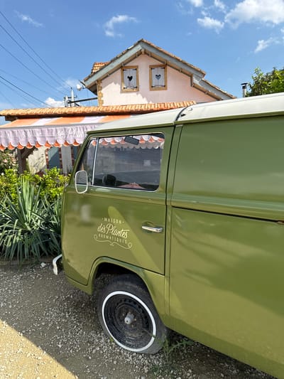 A vintage green van is parked on a gravel and dirt surface in front of a building with a striped awning. The van has a logo on its side that reads "MAISON des PLANTES AROMATIQUES" in white lettering. The building behind the van appears to be a shop or restaurant, with a light pink facade and a tiled roof. There are two windows on the upper story of the building, each with a shutter cut into a heart shape. Lush green foliage and a tall, spiky plant are visible to the left of the van. The sky above is a clear blue with a few white clouds. The scene is bathed in sunlight, casting shadows on the ground. The location is identified as Semogorskiy, Russia.