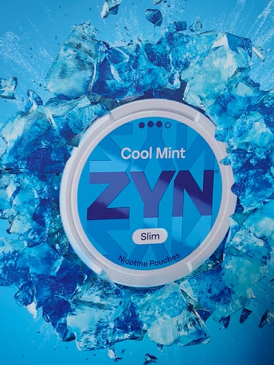 The media file is a close-up, eye-level product shot of a ZYN Cool Mint nicotine pouch container. The container is round with a white rim and a bright blue lid. The lid features the brand name "ZYN" in large, stylized dark blue letters, with a smaller "Cool Mint" label above it and "Slim" in an oval below. The text "Nicotine Pouches" is also visible near the bottom rim.
The container is presented against a vibrant blue background, which is visually fragmented by a dynamic arrangement of shattered ice or crystal-like shapes. These icy elements appear to be exploding outwards from behind the container, creating a sense of motion and a cool, refreshing aesthetic. The ice fragments are various shades of blue, from deep indigo to pale cyan, with sharp, angular edges and some appearing translucent. Small water droplets or ice shards are scattered in the air around the main ice formations.
There are no people visible in the image. The focus is solely on the product and the thematic icy background. The lighting appears bright and even, highlighting the details of the product and the frosty elements. The overall mood conveyed is one of extreme coolness and a crisp, invigorating sensation, aligning with the "Cool Mint" branding. The setting is abstract and studio-like, designed to emphasize the product's flavor profile.