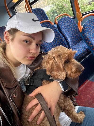 A young woman wearing a white Nike cap and brown leather jacket sits on a blue patterned bus seat with her dog. The dog, a brown Goldendoodle wearing a black harness, is resting on her lap. The woman is also wearing a silver Apple Watch on her left wrist, which is laying on the dog. The bus window is visible in the background, with greenery outside.