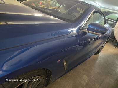 A blue convertible BMW is parked in a garage. The car is predominantly shown from the side, with the front passenger-side quarter panel and door in clear view. The car features an "M" badge on the fender and has chrome trim around the windows. The interior of the car, including a leather seat and steering wheel, is partially visible through the windshield. Other vehicles are parked in the background, along with some construction equipment. The flooring of the garage appears to be concrete. The image is timestamped "21 червня 2026 р. 10:39 PM".