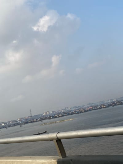 An elevated, slightly tilted view captures a waterfront scene in Ebute-Metta, Nigeria. The upper portion of the frame features a pale blue sky with scattered grey and white cumulus clouds, indicative of an overcast or hazy condition. The lower portion displays a calm, dark blue-grey body of water, likely a lagoon. Along the distant horizon, a dense settlement of stilt houses, constructed from various materials including wood and corrugated iron, extends across the water's surface. Behind these aquatic structures, a land-based urban environment is discernible, featuring multiple buildings and a prominent tall power transmission tower. Atmospheric haze reduces the clarity of distant elements. In the mid-ground on the water, a long, narrow wooden boat is visible, occupied by one individual appearing to operate it. The immediate foreground is dominated by a grey concrete bridge barrier topped with a silver-colored two-bar metallic handrail.