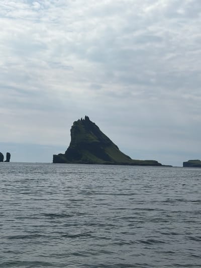 The image depicts a scenic view of the coast near Bøur, Faroe Islands. The foreground is dominated by the sea, with gentle waves creating a textured surface. In the midground, a prominent, dark green, jagged rock formation rises dramatically from the water, its steep slopes covered in vegetation. To the left of this main rock, a smaller, columnar rock formation stands in the sea. Further to the right, a lower, flatter island with grassy slopes is visible. The background consists of a cloudy sky, with soft, diffused light indicating an overcast day. The overall impression is one of natural beauty and rugged coastal landscape.