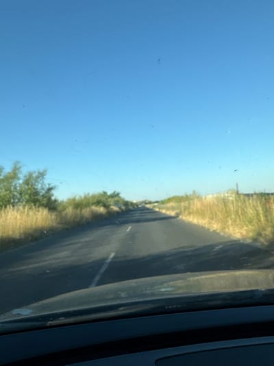 The image shows a view from inside a vehicle, looking out. The perspective is slightly tilted, making the scene appear sideways. On the left side of the frame, a clear blue sky dominates, with a few faint smudges or reflections on the glass.

The right side of the image reveals a roadside scene. A gray asphalt road runs diagonally through the frame, bordered by dry, yellowish grass and some scattered green vegetation, possibly trees or bushes. The lighting suggests it is daytime, likely sunny, given the bright sky and the shadows cast. The scene appears to be a rural or semi-rural area, consistent with a location like Laka, Bulgaria.

The interior of the vehicle is visible on the far right edge, showing a dark, textured surface, possibly part of the dashboard or door panel, with some vertical lines. There are no people or specific objects clearly identifiable as subjects, beyond the road and the surrounding landscape. The overall impression is one of a journey, perhaps on a highway or country road, on a clear, pleasant day. No text is visible in the image.