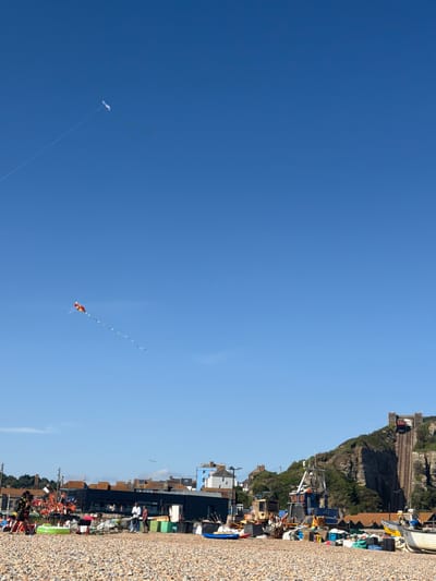 The image depicts a sunny day on a pebble beach in St Leonards, United Kingdom. The sky is a clear blue with two kites visible, one white and one a colorful diamond shape with a long tail. In the background, a row of buildings line the coast, some appearing to be shops or cafes, with darker tiled roofs. A steep, green, tree-covered cliff dominates the right side of the image, with a prominent funicular railway track ascending its face, leading to a building at the top. Scattered on the beach are various boats, fishing equipment, and colorful plastic containers. Several people are visible on the beach, some gathered near boats and others walking. The overall scene suggests a recreational and working coastal environment.