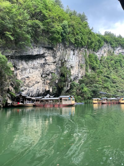 The scene depicts a riverbank in Sanguansi, China. A sheer rock face rises vertically, covered with vegetation, including trees and shrubs. Moored along the water's edge are several multi-story boathouses. The water reflects the structures and the surrounding landscape. The water is colored green.