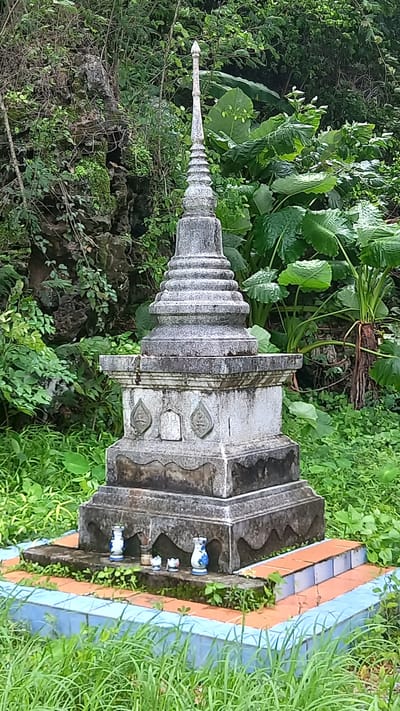 A gray concrete stupa, a Buddhist memorial structure, stands surrounded by green vegetation in Thap Put, Thailand. The stupa is multi-tiered with a pointed spire and decorated with leaf-like designs. Miniature vases and figurines are placed around the base. The stupa sits within a tiled border painted in blue and orange. The surrounding greenery includes grass, small leafy plants, and larger-leaved plants.