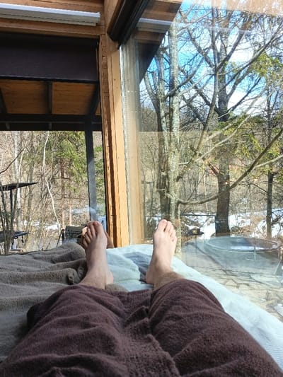 The image depicts a first-person perspective from an indoor setting, looking outward through a large window, located in Korostiv, Ukraine. In the foreground, the bare feet and lower legs of an individual are visible, resting on a light blue-grey blanket. The individual is covered from the waist down by a dark brown, textured blanket or robe. The interior features light brown wooden window frames and structural elements; a dark roller blind and a white light fixture are visible in the upper left. The exterior view through the window reveals a winter or early spring landscape under a clear blue sky. Dominant features include deciduous trees with bare branches and some coniferous trees, with patches of snow covering the ground. To the left of the exterior scene, a dark metal structure is partially visible. To the right, an oval-shaped dark outdoor hot tub or plunge pool rests on a stone patio, partially snow-covered, with a small table positioned beside it.
