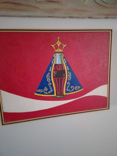 The image displays a framed piece of artwork mounted on a light gray wall in Campo Grande, Brazil. The artwork features a stylized depiction of a Coca-Cola bottle transformed into a religious figure, likely a Marian image. The bottle, rendered in green glass with a red label partially visible as "Coca-Cola" in black, is adorned with a golden crown topped by a cross. A blue mantle, decorated with intricate golden and white scrollwork, drapes over the bottle, extending to its base. The background is predominantly red, incorporating a curved white stripe and a lower red stripe, emblematic of the Coca-Cola brand's visual identity. The artwork is enclosed within a thin, light-colored frame with a black inner border. Above the frame, a textured portion of another object or artwork, exhibiting earthy tones of brown, green, and light blue, is partially visible.