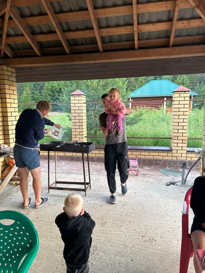 In the city of Pepelyaeva, Russia, a man in a dark blue jacket and blue shorts is standing by a black barbecue grill, holding a clear plastic bag and appearing to be adding something to the grill. He is wearing blue Croc-style sandals. To his right, another man in a grey t-shirt and black pants is holding a young child wearing a pink sweater and pink pants. The child is looking towards the camera. In the foreground, a young boy with blond hair, wearing a black hoodie, is facing away from the camera, with his hands to his ears. The scene takes place outdoors under a wooden roof structure with a metal roof. Behind the barbecue, there is a brick wall and a natural landscape with trees and grass, and a building with a teal roof. There are also several chairs visible, including a green plastic chair in the lower left and a red chair on the right. The ground is paved with concrete.