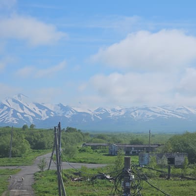 The image depicts a serene landscape in Sokoch, Russia, likely during daytime under clear skies with scattered clouds. The scene is dominated by a majestic snow-capped mountain range, partially obscured by a thin layer of mist or clouds, evoking a sense of tranquil grandeur. In the foreground, an abandoned, weathered structure, possibly a building or industrial remnant, stands amidst lush green vegetation and overgrown grass, hinting at a forgotten past or a remote location. A paved road winds through the scene, leading the eye towards the distant mountains.

Prominently featured are a series of utility poles with a tangled network of electrical wires, suggesting the presence of infrastructure in this otherwise remote setting. The poles are weathered and some of the structures they support appear dilapidated, further reinforcing the theme of abandonment or decay. The surrounding environment is characterized by abundant greenery, with trees and bushes creating a vibrant contrast to the muted tones of the mountains and the decaying man-made structures.

The overall mood of the image is one of quiet solitude and natural beauty, with a touch of melancholy due to the presence of abandoned structures. The natural elements, the snow-capped mountains, and the vast sky convey a sense of scale and permanence, while the man-made elements speak of human presence and its ephemeral nature. There is no visible text in the image.